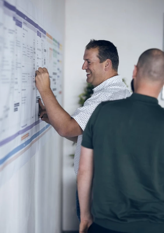 Two professionals evaluating a project timeline on a wall-mounted board in an office setting.